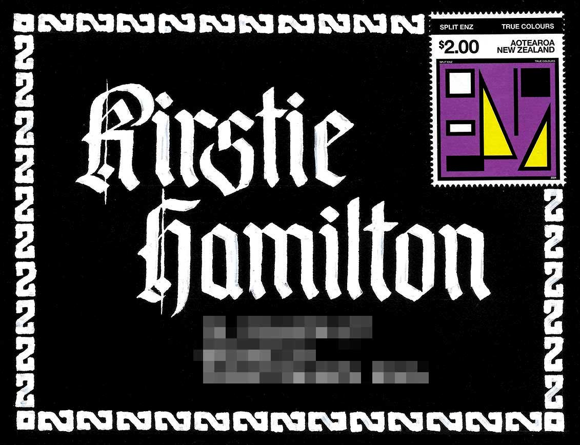 A black envelope addressed to Kirstie Hamilton in large blackletter calligraphy. The name and address are written in white ink, with a white patterned border around the outer edge.